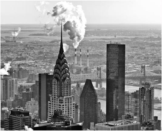 Monochrome picture of the Chrysler building. It is taken from a high view point. There are other tall buildings nearby. Just beyond is a three tall chimney industrial plant belching smoke or steam into the early morning sky.
