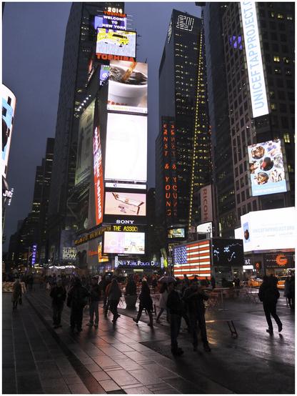 A night shot of the bright neon lights of Times Square. There are many tourists in the foreground.