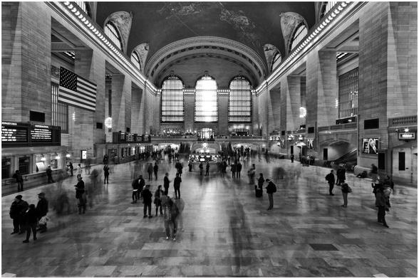 A monochrome image of Grand Central Terminal. There are columns either side of the image supporting a high arched roof. Centre, back of image are three large arched windows.. there are many people on the concourse.