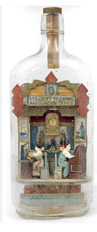 Photo of a large glass bottle. Inside is a scene in Berghoff Brewing Association Office featuring a man and woman working back to back in booths. On the walls are pictures and in the centre a large clock.