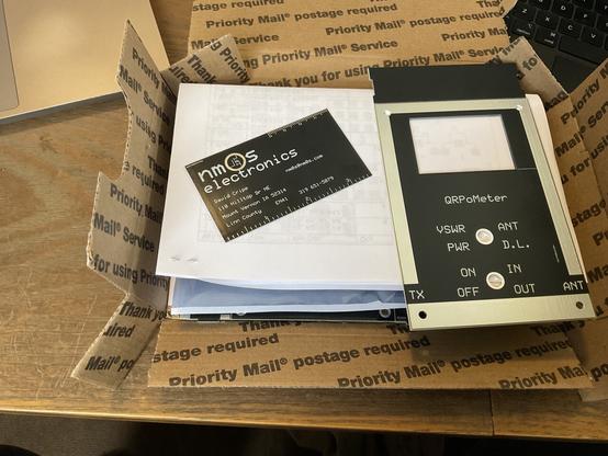 On the left a black single layer printed circuit board, the size and shape of a standard business card, showing the logo and contact information for "NM0S Electronics."

On the right, a black multi-layer printed circuit board with the word "QRPoMeter" and several abbreviations silk-screened in white.

Both are sitting on top of remaining parts and printed instructions, inside of an unfolded US Postal Service Priority Mail box.