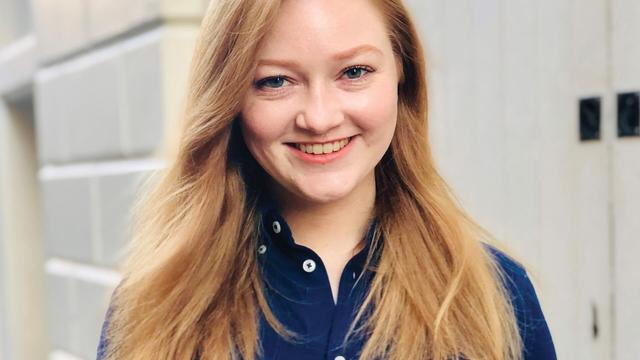 A F Steadman smiling at the camera. She is wearing a dark blue shirt and is in front of a white door and a white brick wall.