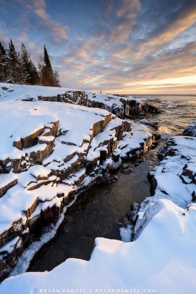 A photo of a snow-covered basalt shoreline under linear clouds painted yellow from the sun which is off camera right.