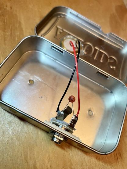 Photo of an open Altoids tin with a guitar jack and piezo disk installed. Also a capacitor connected to the guitar jack.