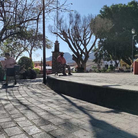 En la plaza central de Tepoztlán, un hombre sentado en una silla, confundido entre las sombras de árboles y construcciones.