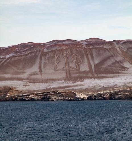 A view from sea looking back to shore where the land rises quickly to form a shallow-jagged dirt hill that dominates the central third of the shot. The sea in the bottom third is generally calm. The sky is filled with thin cloud practically whiting out the view above. The notable aspect of the pale and mid brown landscape is the prominent shape marked on its surface. It resembles a candelabra with three spokes. The outer two further branch twice up their lengths into two more spokes, as the taller central spoke does once at its peak.