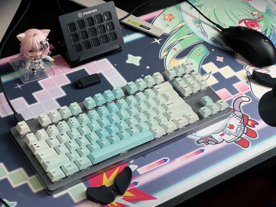 A photograph of my desk space! The desk itself is a dark brown wood (almost black). On the desk is a Nyanners nendoroid. Beside it is the Elgato Stream Deck (black variant). My keyboard is metallic silver with a gradient blue keycap set decorating it. I use the Logitech G703 wireless as my mouse to the right of the keyboard. My two DeltaHub Carpio 2.0 wrist rests (black) can be seen.