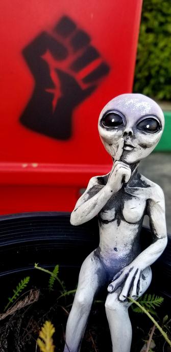 My 2nd alien figurine, making a be quiet sign with hand, sitting in a plant pot, with black activism fist painted on a red newspaper box.