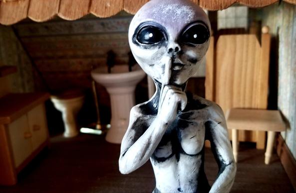 My alien figurine, making a shush sign with hand, sitting partway in my dollhouse bathroom.