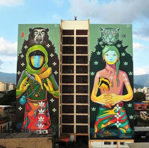 Streetartwall. Huge mural on a skyscraper of about 10 floors. In the middle is a double row of windows. On each of the two windowless side walls are sprayed / painted two girls in naive style. The left girl wears a green headscarf, green sweater and red skirt. Her face is painted in three stripes in color. She holds her arms upwards at the level of her chest. Behind her appears a bear, like a shadow.
The second girl has a tiger as a shadow. She is wearing a colorful skirt, pink shirt and a braid. Her arms are folded in front of her.
