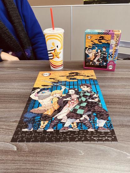 A completed jigsaw puzzle of the anime Kimetsu no Yaiba with my wife, a Hardee’s cup of pop, and the puzzle box in the background. 

The puzzle picture shows the four heroes of the anime brandishing blades.