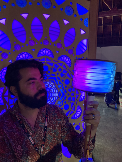 Dimly lit bearded guy holds a spinning light box that blurs in the foreground. He stands in front of a Purplish Glowing led mandala lit in harmony with the cube.
