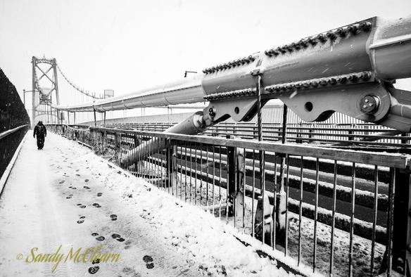 Man walking toward camera on the snow covered walking path of a suspension bridge.