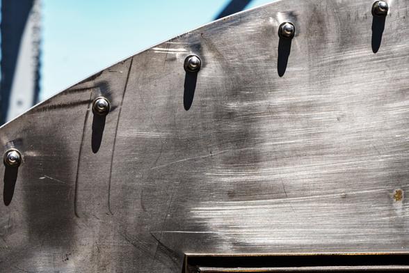 A photograph of a portion of the steel cover over the gear mechanism behind the main operating wheel on one side of a bascule bridge. The cover fills most of the frame, the metal is scratched, and bolts protrude along the facing edge at regular intervals, the sun casting their shadows downward along the cover. A pale blue sky is visible in the top left of the frame.