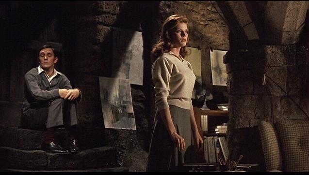 A man sitting on the table with his hands arounfmd his knees in the background. A woman stands facing forward.