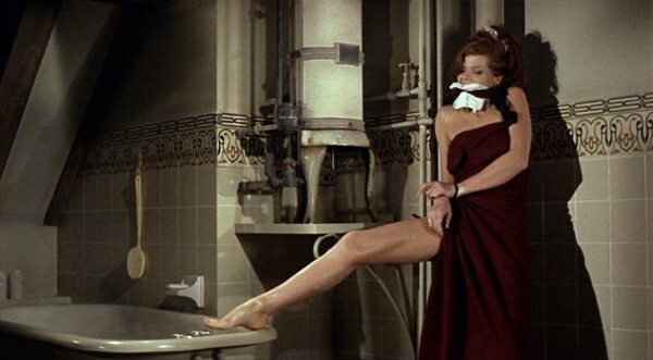 A woman wrapped in red bath towel and gagged, tied to a post, trying to get to a razor placed on the edge of the bathtub with her foot.