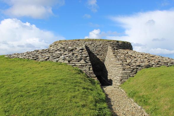 The outside of the tomb. The entrance passage is now partly open. It's quite tidy looking drystone walling, set in green grass. Blue sky, sunshine. A gravel path leads towards the passage.
