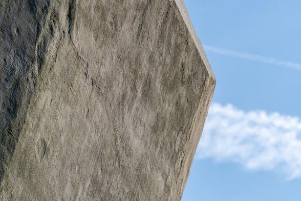 A color photograph of a giant concrete block serving as a counterweight for a bascule bridge. Zoomed in on a corner,  there are clouds and a blue sky behind.