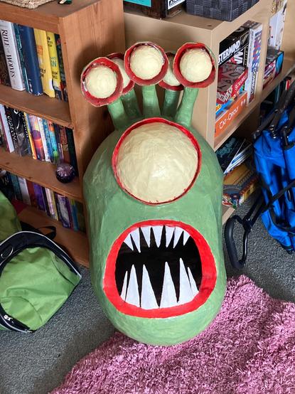 A painted cardboard and paper mache sculpture of a Dungeons & Dragons monster, the Beyonder, with a green oval head, an open mouth with red gums and long, sharp white teeth on the bottom half of the face, one large yellow eye with red rims on the top half of the face, and five smaller eyes on stalks emerging from the top of the head