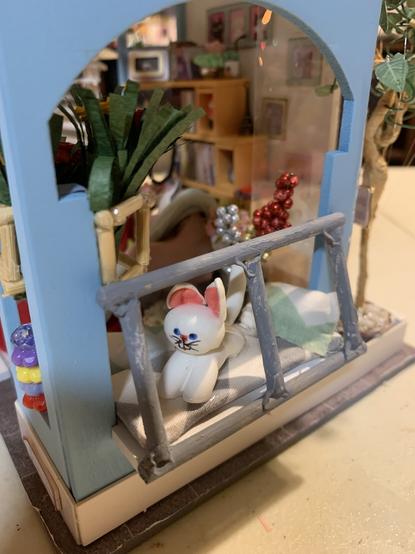 Close up on the face of the white cat with pink ears made of clay standing in on the gray and white bench cushion in the bay window