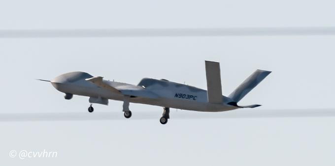 an all grey unmanned aerial vehicle is sandwiched between two power lines. it has a bulbous nose and underneath is a sensor turret. Its landing gear is down just behind the thin wings on the top is a intake for the engine. It has a separate slab V tail and the exhaust is oval shaped and partially flat. While this is clearly a military vehicle it cares a civilian registration  stenciled on the rear fuselage