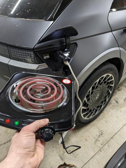 A portable coil top stove visibly glowing, the stove is plugged into a power strip which is plugged into the charge port of a Hyundai Ioniq 5