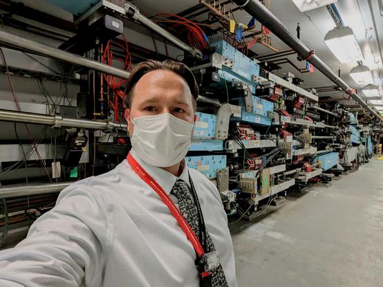 A masked man and a shirt and tie wearing a dosimeter around a lanyard stands in front of a stack of beamlines full of magnets.