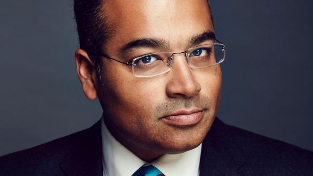 Krishnan Guru-Murthy looking at the camera. He is wearing a black suit and a white shirt. He is in front of a dark grey background.