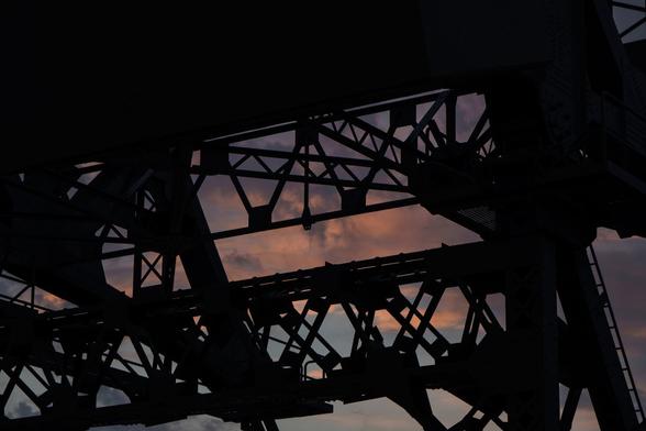 A picture of the shadowed upper structure of a bascule bridge. Behind the silhouetted girders, the early morning sky is a darker blue, with orange/purple clouds.