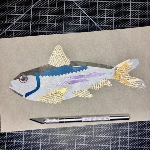 A multicolored alewife fish collage in silver with dark gray, teal, gold, and torn bits from an antique book of poems for the fins.