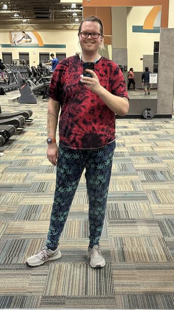 Me standing upright looking at a mirror in the gym wearing a red tie dye t shirt and pants with a weed leaf print on them looking at the camera on my phone smiling.