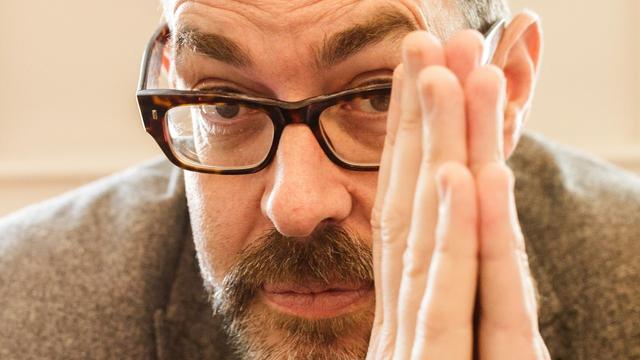 Close-up of Richard Osman looking at the camera. He is wearing rectangular brown glasses and has his hands palms together just in front of his face. Photo credit: Conor O'Leary