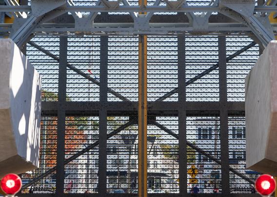 A view through the metal grid of a bascule bridge’s deck in the raised position. Red warning lights are in the lower corners of the frame, and the buildings on the other side of the river are visible through the desk.