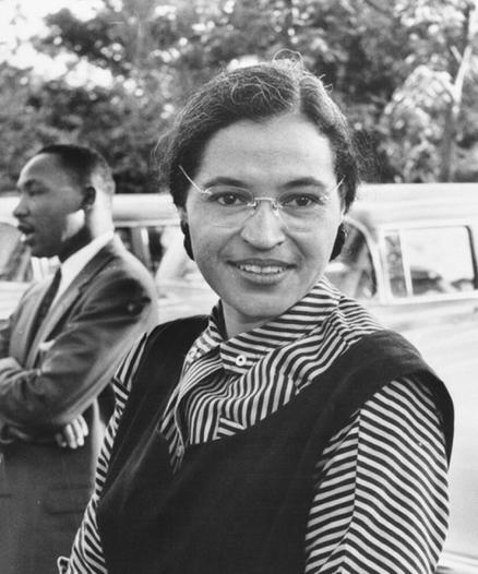 Black and white photo of Rosa Parks. Outdoor shot of a light skinned Black woman wearing wire-rimmed glasses with hair pulled back. She has on a bold striped blouse with a dark sleeveless dress over it. She’s smiling and standing in front of a car. 

The Rev Dr Martin Luther King, Jr. stands in the background. He is a Black man with a mustache and short hair. He wears a darks suit and tie, has his arms crossed, and looks to be in mid conversation with someone out of frame.