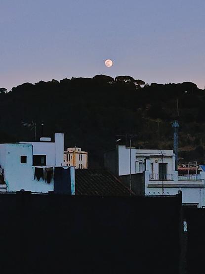 Lluna plena sobre un turonet allargassat. Davant, en primer pla, és veuen les part del darrera d'unes cases velles de poble, color blanc. El cel blau grisós, amb la lluna i les cases blanques contrasten amb la negror de la silueta de la muntanya.