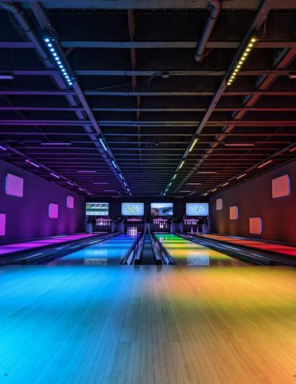 Four bowling alleys with led lightning bars putting entire scenery in different colors. The perpective is straight from the center and slightly from bottom up. There aren't any people at this moment. The dark ceiling with some technical ingredients is a stark contrast to the light parquet on the ground.