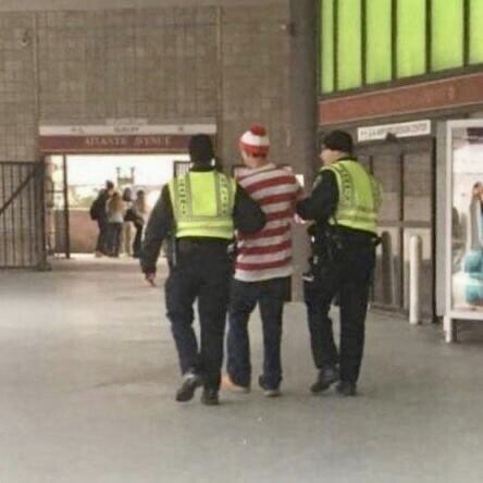 Man in Where’s Waldo costume being detained by 2 members of law enforcement.