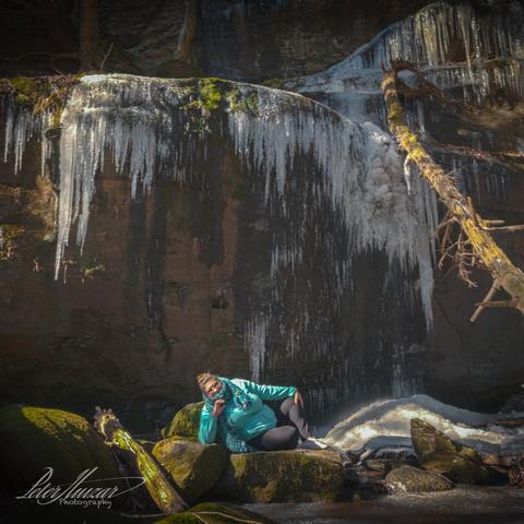 Woman sitting in front of rock cliff with icicles melting off it. Dead tree on right.