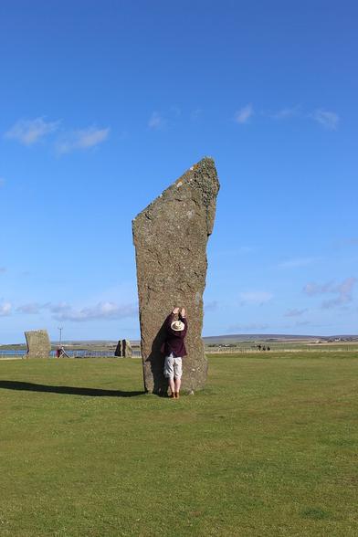 A white man in shorts and a jacket, wearing a straw hat, leans against a standing stone, stretching his arms up as high as he can. He reaches about a third of the way up. The stone is narrower at the bottom and widens towards the top, which is steeply angled. Other, shorter stones are visible in the distance. The sky is very blue, and the sun is shining.