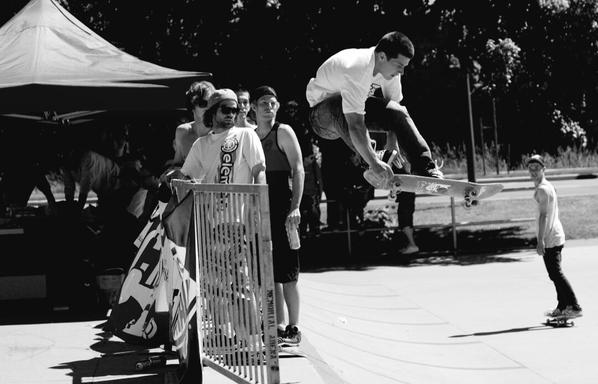 Black & white photo. Skateboarder in white tshirt & denim jeans, performing a blaster aerial move, over a metal gate.