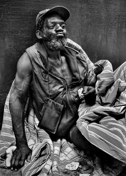 A Black homeless US veteran sirs on the concrete with a blanket.