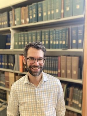 A smiling man wearing tortoise-shell glasses and an open collar checked pattern shirt. He has slightly graying hair, beard, and mustache. He has a high, academic forehead. He is standing in front of a large shelf of bound volumes of books in a library. He looks like he would be happy to help you find the book you're looking for.