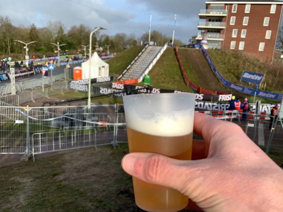 In the foreground, a hand raised with a glass of beer, with the steep descent and stairs of the Hoogerheide cyclo-cross course in the background.