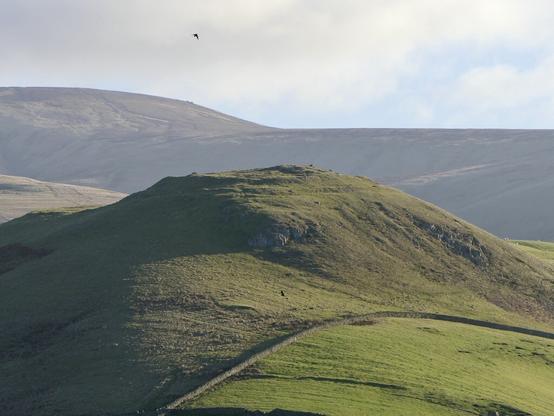 Rocky hill top with ramparts around a flat summit lit from the right with smooth rolling hills in the background.