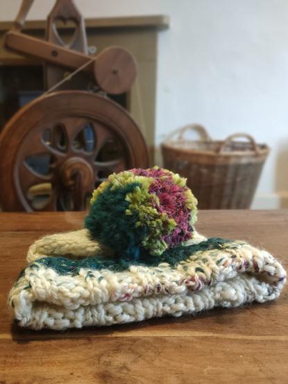 Bobble hat on wooden table. Viewed from the folded edge, w bobble folded over. Mallard green, lime green and pink bobble. W spinning wheel and wicker basket in background.