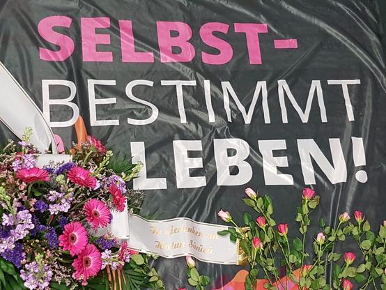 Bild zeigt einen Blumenkranz mit Schleife „zum Gedenken an Hatun Sürücü“ auf einer Fahne. Die Fahne trägt den Schriftzug „Selbstbestimmt leben!“