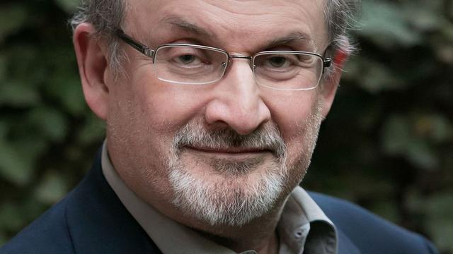 Close-up of Salman Rushdie looking at the camera. He is wearing rectangular glasses and is in front of green leaves. Photo credit: Beowulf Sheehan