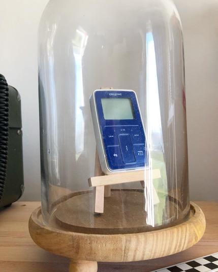 A small, handheld MP3 player – about the size of a compact cassette – displayed inside a glass vitrine. The face of the MP3 player is deep blue, while its back is bright white.