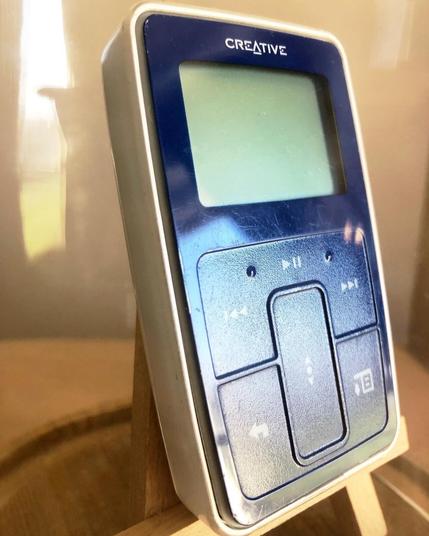A small, handheld MP3 player – about the size of a compact cassette – displayed inside a glass vitrine. The face of the MP3 player is deep blue, while its back is bright white. This extreme closeup shows that the device's buttons have a slightly grainy texture, while the rest of the front face is smooth (yet shows scratches from wear and tear).