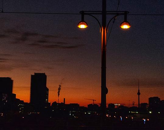Blick von der Warschauer Brücke über Berlin, im Vordergrund eine Straßenlaterne, im Hintergrund ein Hochhaus, rauchende Schornsteine, ein Kran und der Fernsehturm. Der Himmel ist rot und orange, die Gebäude sind nur als schwarze Schatten/Silhouetten zu erkennen.